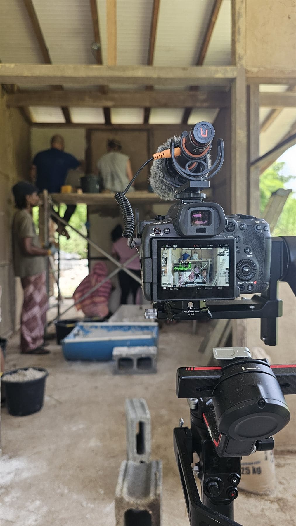 A cinema camera with a shotgun microphone on a gimbal records an earthen-building crew working on scaffolding inside a timber frame; its screen shows the live picture and audio levels, the crew soft behind it.