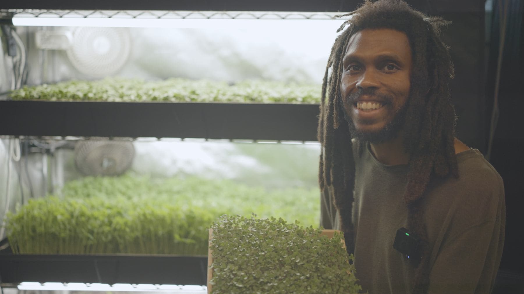 A grower with long locs smiles beside racks of microgreens under bright tube lights and circulation fans, holding a tray, a wireless lapel microphone clipped to his shirt.
