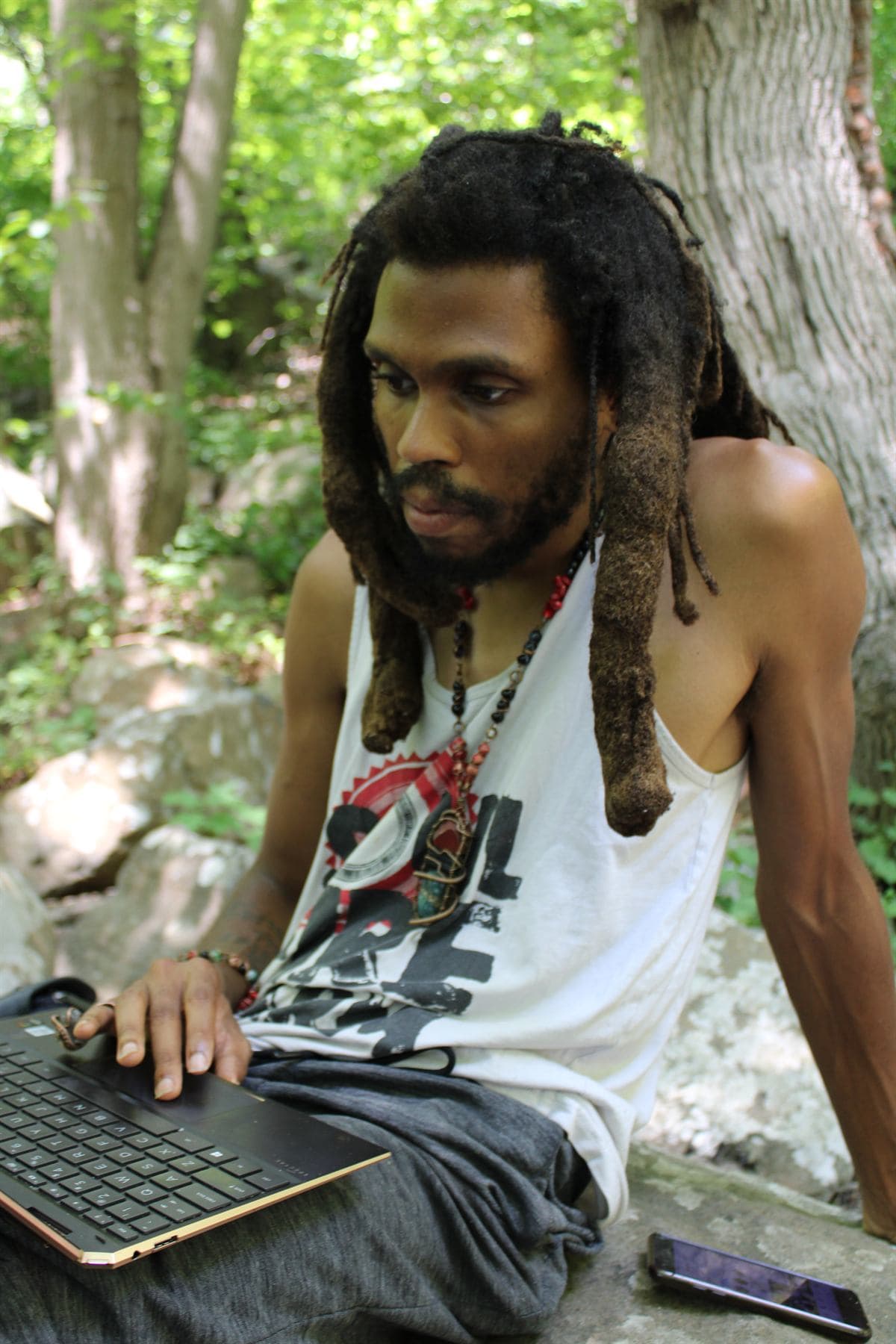 A man with long locs sits on a rock in the woods with a laptop open across his lap, one hand on the trackpad, reading the screen, a phone lying on the rock beside him.