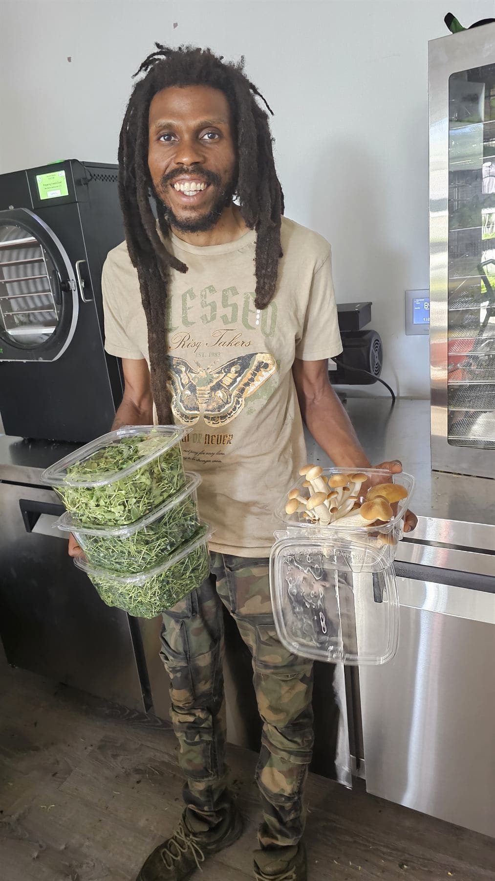 A grower grins while holding stacked clamshells of microgreens and a clamshell of mushrooms at a steel prep counter, a freeze dryer with a lit status screen and a commercial dehydrator behind him.
