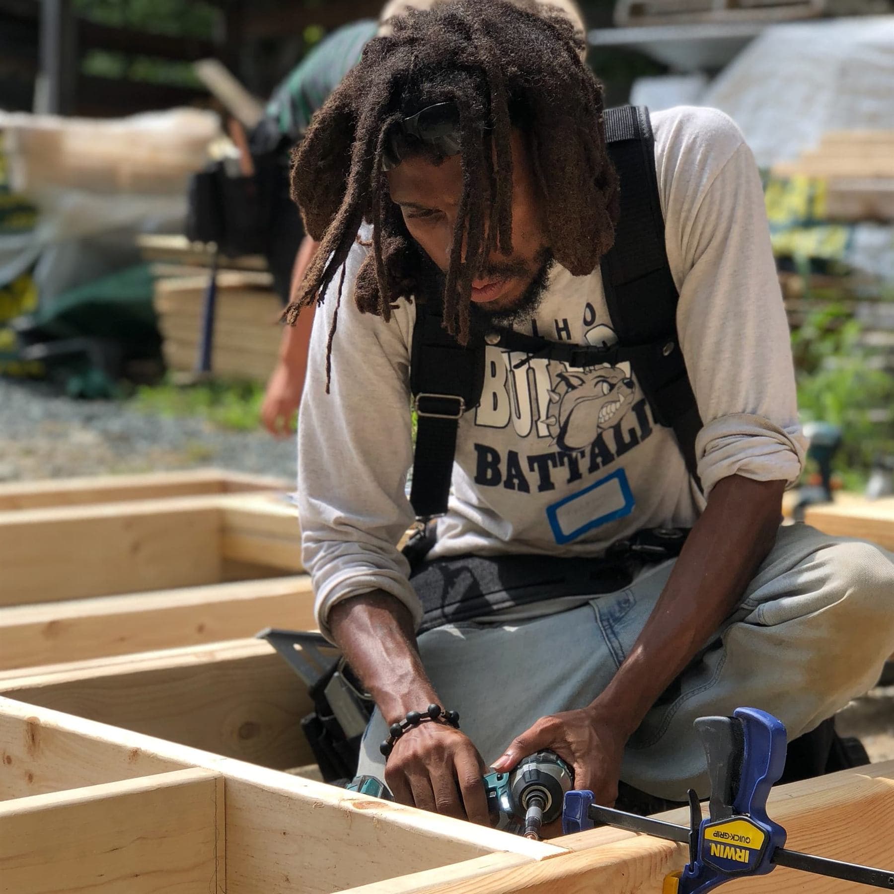 A man with long locs and safety glasses pushed up on his head kneels in a tool harness, driving a screw with a cordless impact driver into a raised-bed frame held with a quick-grip clamp.