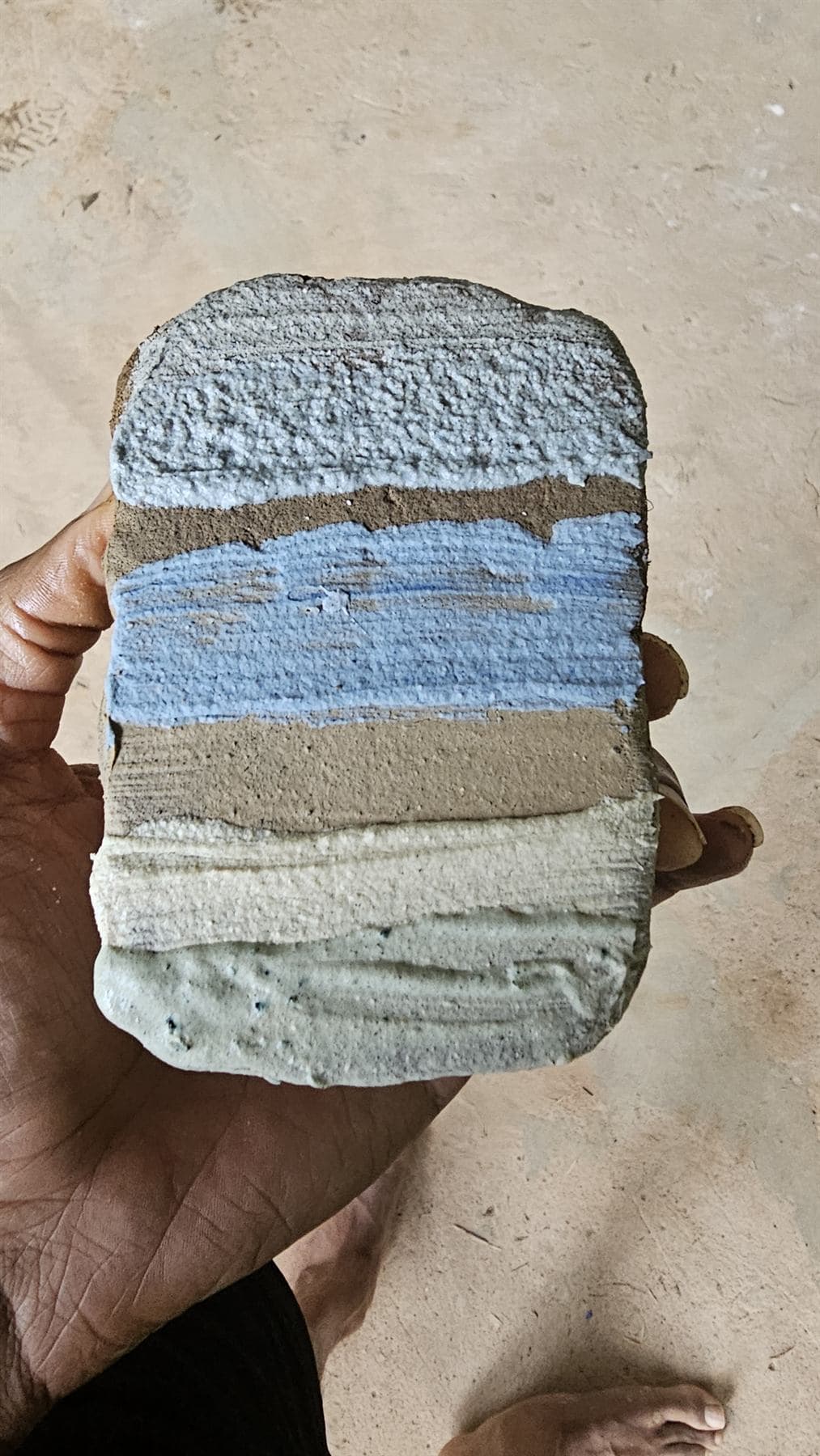 An open hand holds up a small slab built from layered bands of textured material — pale grey, brown, blue, brown, cream and sage green — over a sandy concrete floor, a bare foot at the edge of the frame.