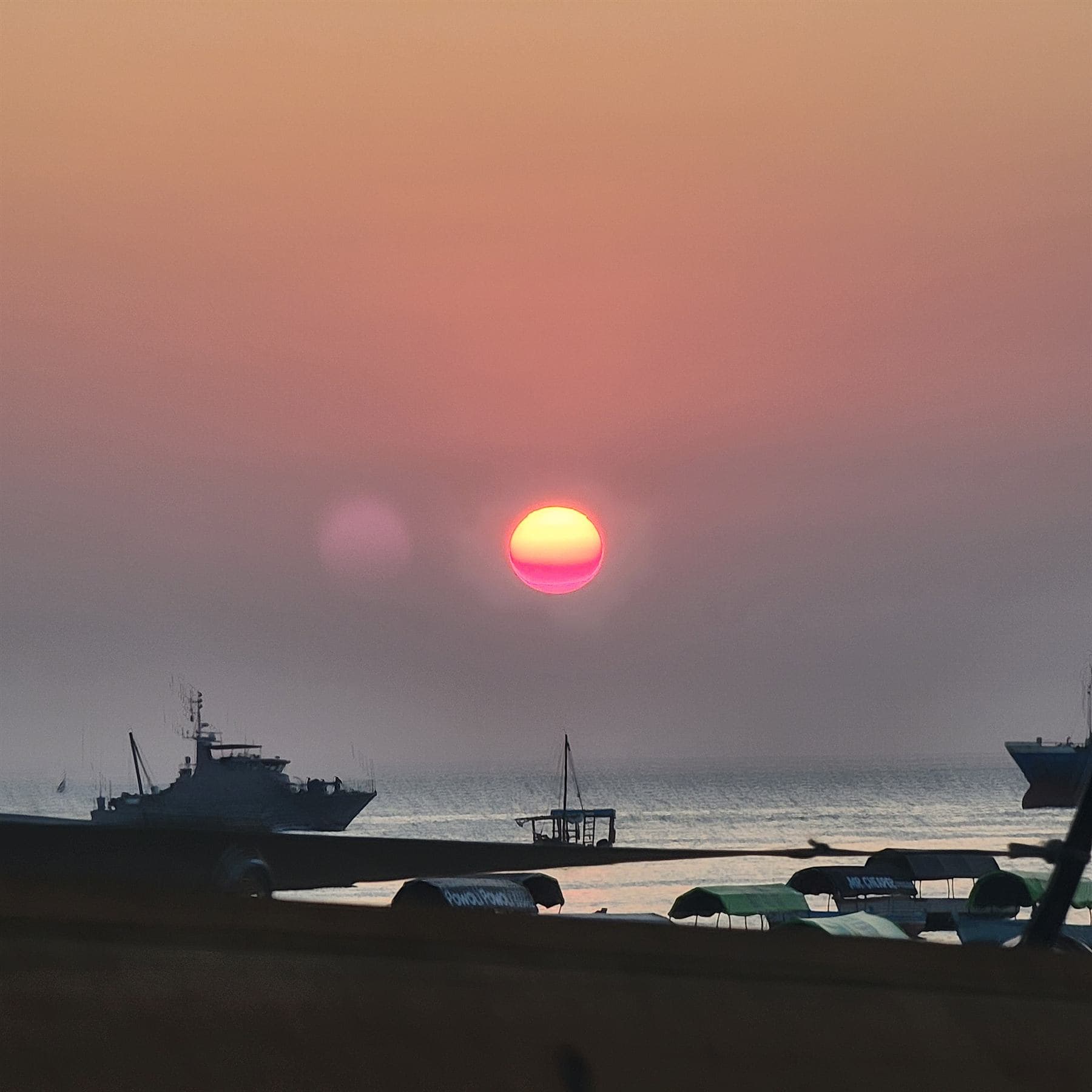 A red sun low in a hazy pink sky over a harbour, a patrol vessel, a small wooden boat and a cargo ship in silhouette, canopied tour boats pulled up in the foreground.