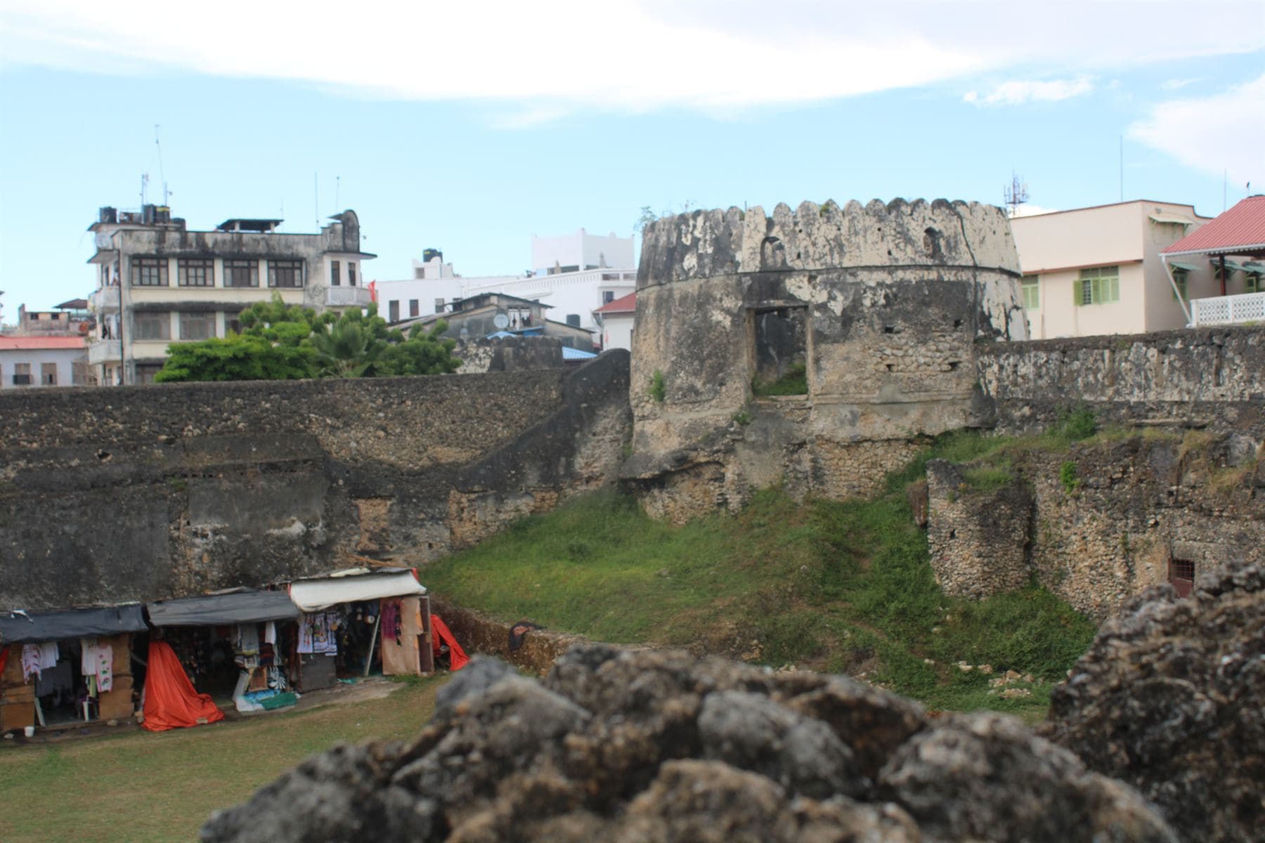 The weathered coral-stone walls and round crenellated tower of an old fort, grass inside the walls, a row of small market stalls hung with cloth at lower left and apartment buildings beyond.