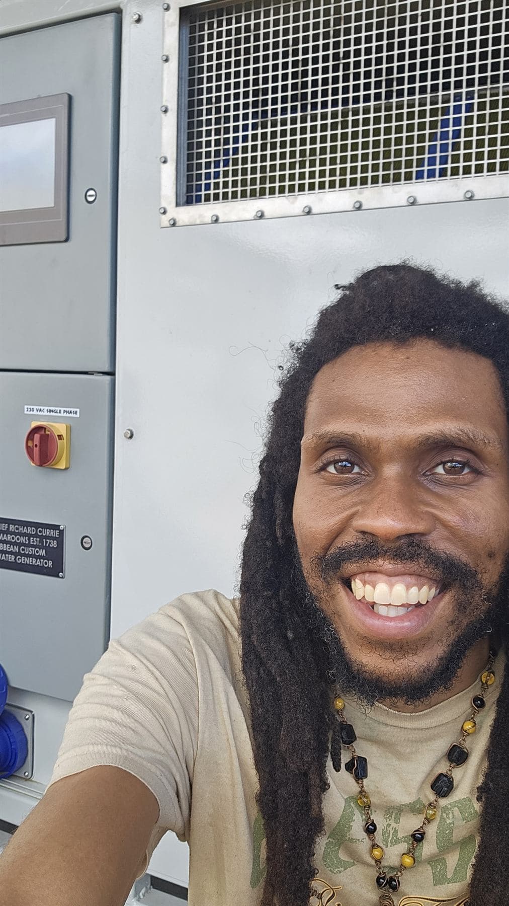 A smiling man with long locs takes a selfie beside a large atmospheric water generator, its control screen, air grille, 220-volt switch and nameplate visible behind him.